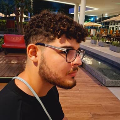 Profile photo of João Gabriel, a young man with curly hair, trimmed beard, and large glasses, sitting outdoors near a water feature at night.